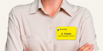 En person bär en ljusbeige skjorta med knappar och lätt uppvikta ärmar. Personen har armarna i kors. På vänster sida av bröstet finns en gul namnbricka med en blå och svart logotyp, texten 'Technology', namnet 'E. Platier' och titeln 'RESPONSABLE ACHATS' i svart text. Neutral bakgrund för tydlighet.