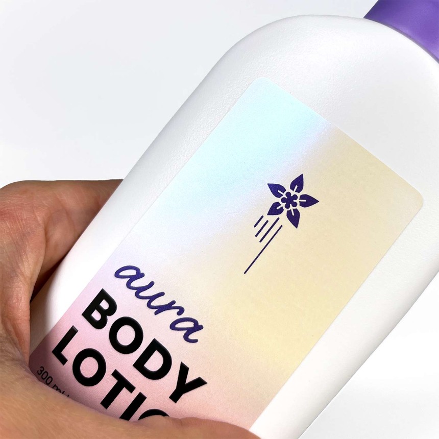 Etikett i pärlemorsskimrande folie med texten "aura body lotion". Här syns den skimrande effekten på en vit flaska.