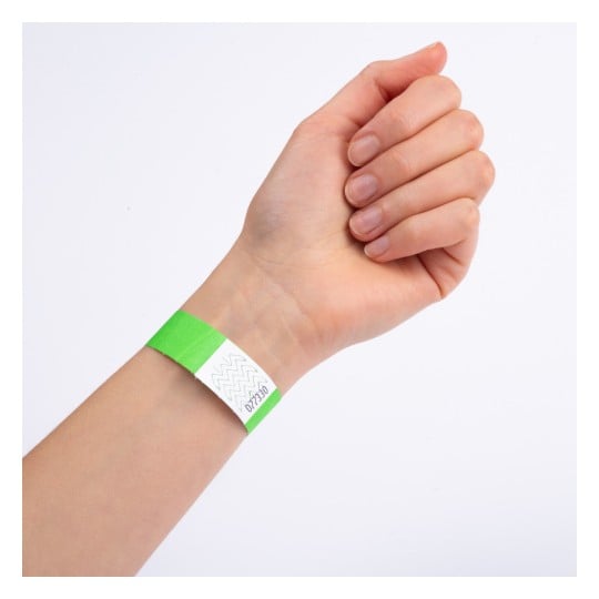En närbild av en hand med ett grönt armband på handleden. Armbandet har en vit sektion med ett sicksackmönster och verkar vara märkt med ett nummer.