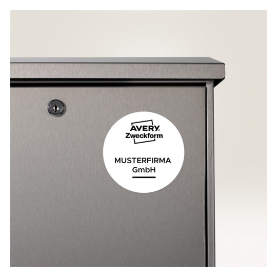 Bilden visar en närbild av ett grått metallföremål med ett nyckelhål och en cirkulär etikett. Etiketten har texten 'AVERY Zweckform MUSTERFIRMA GmbH', vilket tyder på att det kan vara en förvaringsenhet eller ett skåp.