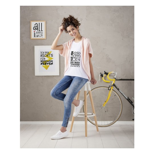 Bilden visar en ung kvinna med lockigt hår som sitter på en pall, iklädd en vit t-shirt med ett uttalande, en ljusrosa kofta och blå jeans. I bakgrunden finns två inramade tryck och en cykel.
