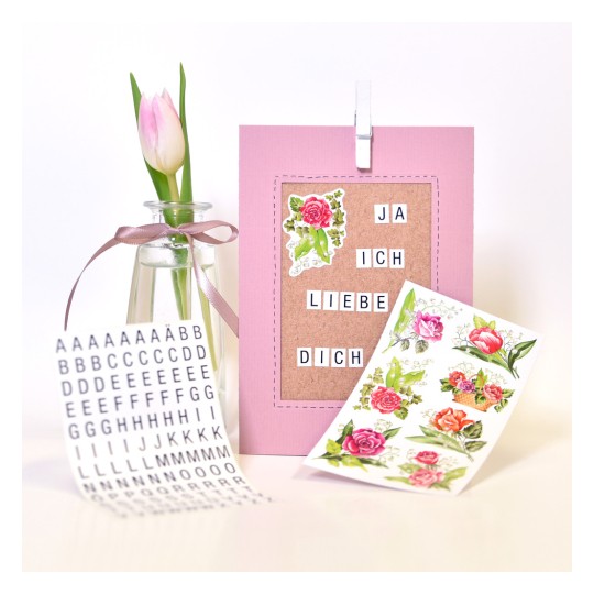 Bilden visar ett rosa kort med texten 'JA ICH LIEBE DICH' (Jag älskar dig), dekorativa blomklistermärken, en liten vas med en tulpan och ett ark med alfabetklistermärken.