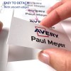 Bilden visar en hand som lossar en etikett från ett ark, med namnet 'Paul Meyer' och 'Avery'-logotypen, med en anteckning som indikerar att den är lätt att lossna med släta kanter.