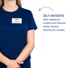Bilden visar en person som bär en mörkblå skjorta med en namnbricka där det står 'B. Carlos, Marknadsföringschef.' Dessutom finns det text som beskriver namnbrickets självhäftande egenskap, och som säger att den sitter fast pålitligt på kläder och kan tas bort enkelt utan att lämna några rester.