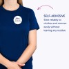 Bilden visar en person som bär en blå t-shirt med en självhäftande namnskylt där det står 'B. Carlos, Marknadschef.' Texten understryker att skylten fäster pålitligt på kläder och kan tas bort enkelt utan att lämna kvar några rester.