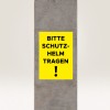 Bilden visar en gul skylt på en betongpelare med texten 'BITTE SCHUTZ-HELM TRAGEN!' vilket översätts till 'Var vänlig bär hjälm!' och anger ett säkerhetskrav.