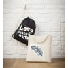 Bilden visar två väskor mot en vit tegelvägg. En svart dragskoväska med orden 'Love, Peace, Party' hänger. Bredvid den visas en beige tygväska med en dekorativ fjäderdesign.